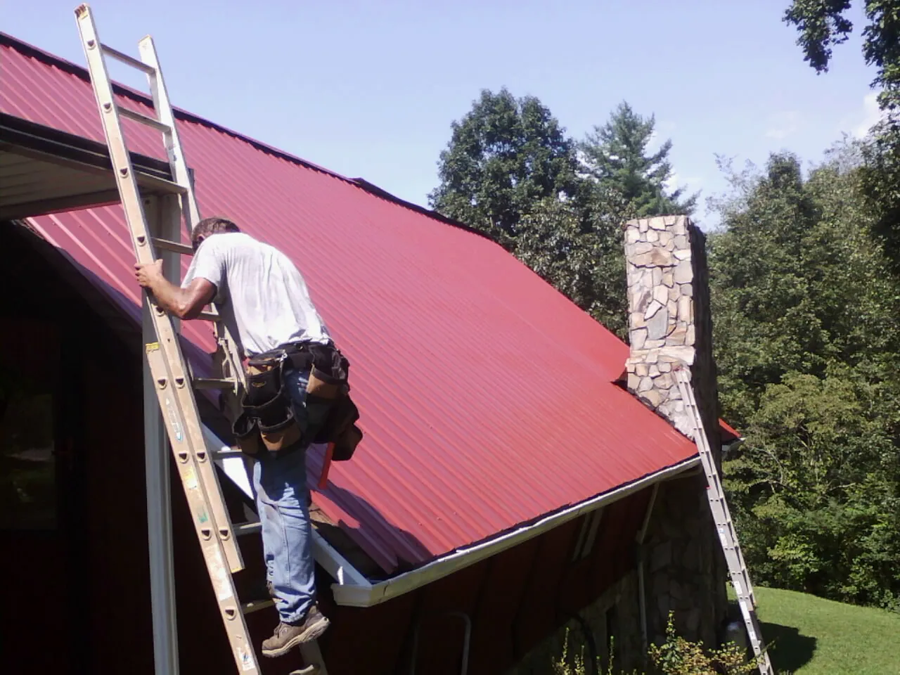 Roofer on a ladder beside a red metal roof and stone chimney — Soffit & Fascia Repair by Roofing Brothers in Ecorse